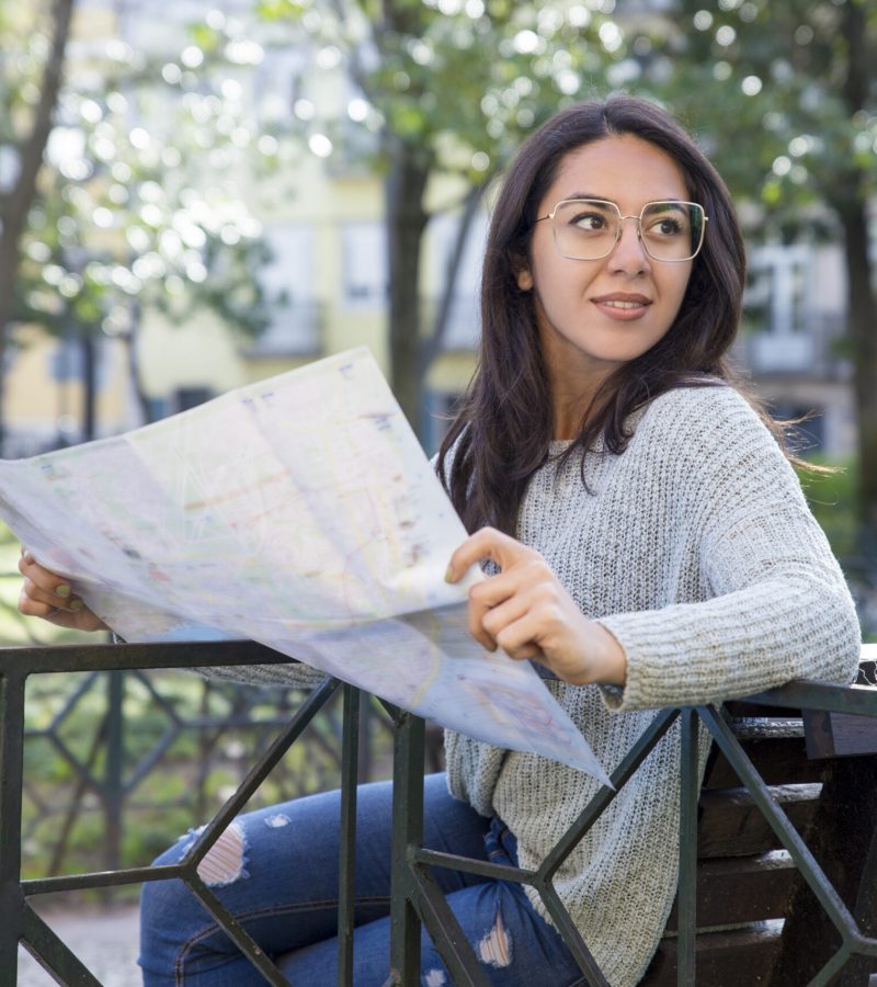 Content pretty young woman using paper map on bench outdoors. Beautiful lady sitting on bench and searching for city sights with blurred trees and buildings in background. City trip concept.